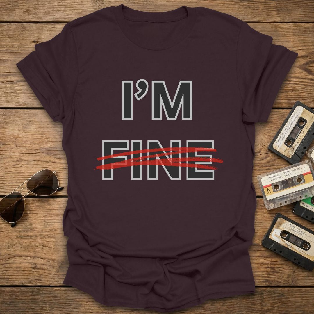 Brown t-shirt with 'I'm Fine' with fine crossed out in red text on a wooden surface with sunglasses and cassettes.