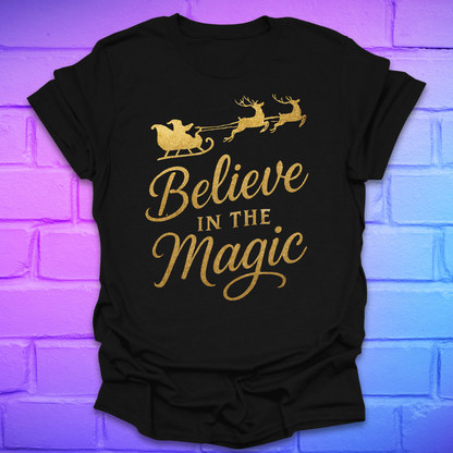 Black t-shirt with gold 'Believe in the Magic' text and Santa sleigh design on a brick wall background.