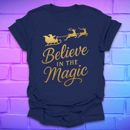 Navy blue t-shirt with gold 'Believe in the Magic' text and Santa sleigh design on a brick wall background.