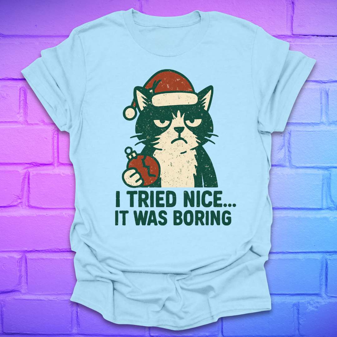 Light blue t-shirt with a frowning cat holding a broken christmas ball with text, 'i tried nice. it was boring'. on a brick background.