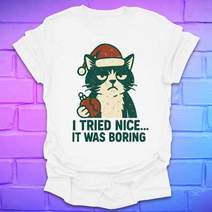 White t-shirt with a frowning cat holding a broken christmas ball with text, 'i tried nice. it was boring'. on a brick background.