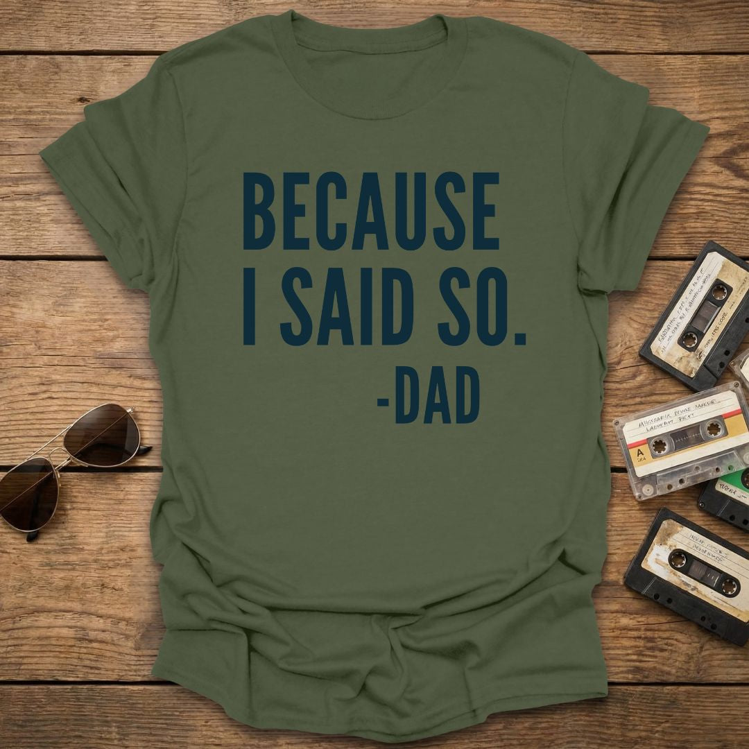 Green t-shirt with 'BECAUSE I SAID SO -DAD' text on a wooden surface with sunglasses and cassettes.