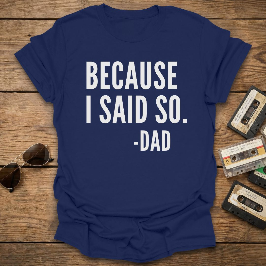 Blue t-shirt with 'BECAUSE I SAID SO. -DAD' text on a wooden surface with sunglasses and cassettes.