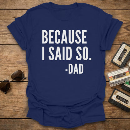Blue t-shirt with 'BECAUSE I SAID SO. -DAD' text on a wooden surface with sunglasses and cassettes.