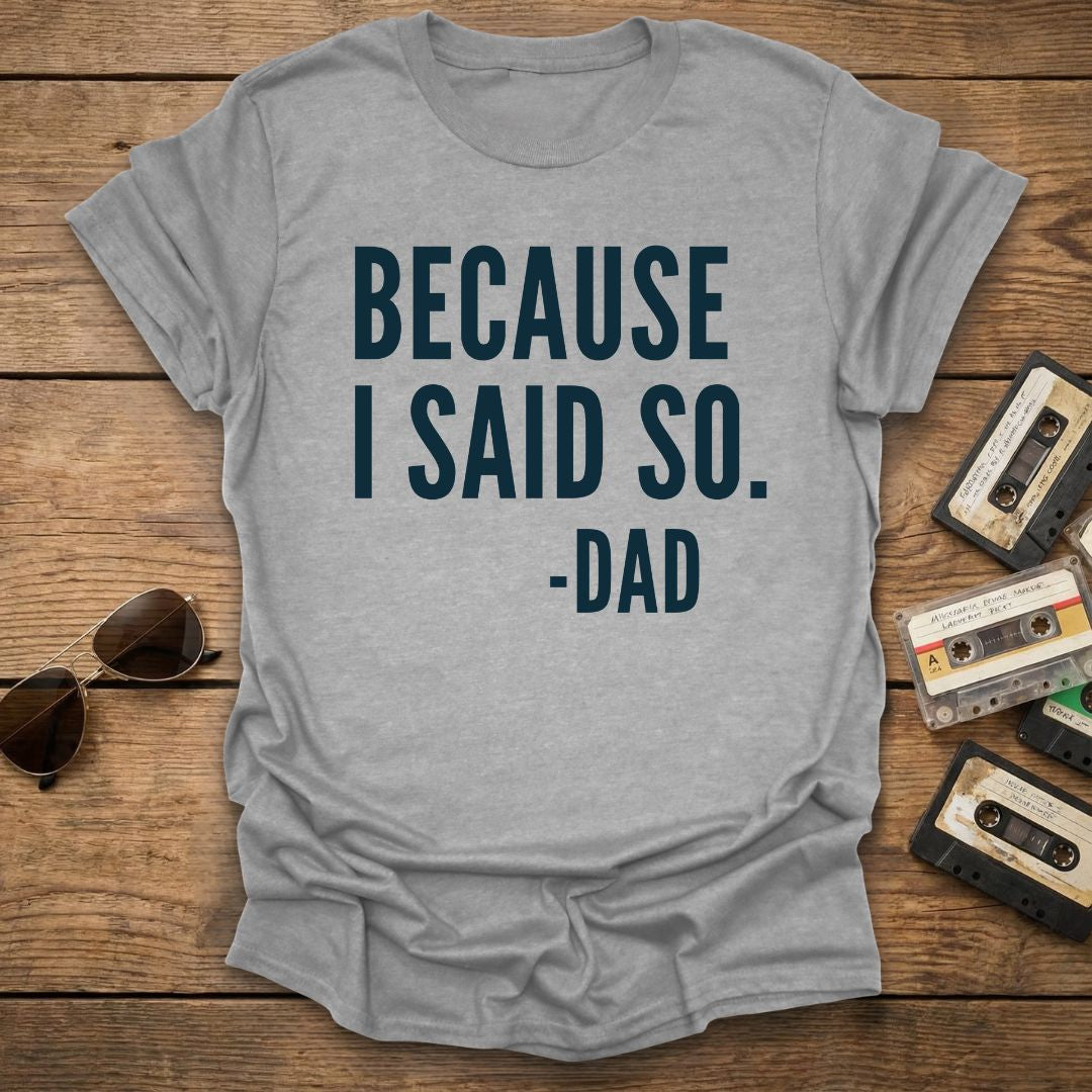 Gray t-shirt with 'BECAUSE I SAID SO. -DAD' text on a wooden surface with sunglasses and cassettes.