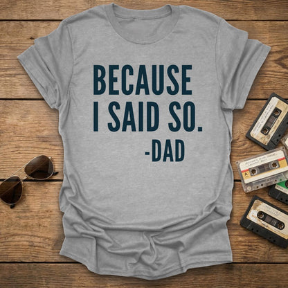 Gray t-shirt with 'BECAUSE I SAID SO. -DAD' text on a wooden surface with sunglasses and cassettes.