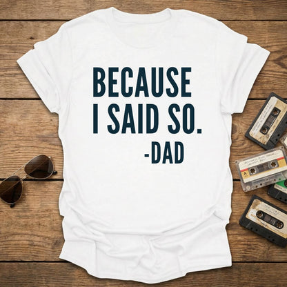 White t-shirt with 'BECAUSE I SAID SO. -DAD' text on a wooden surface with sunglasses and cassettes.