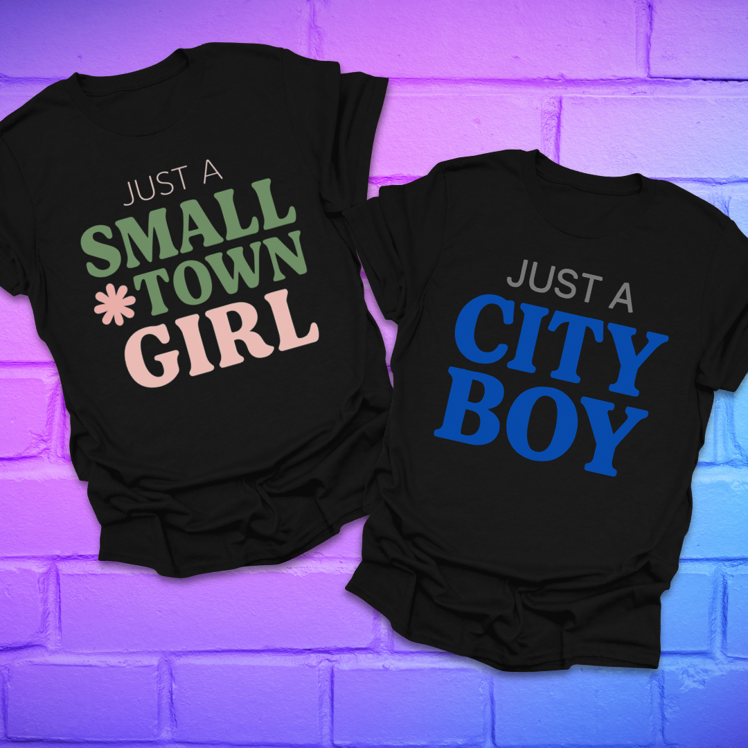 Two black t-shirts with 'Just a Small Town Girl' and 'Just a City Boy' text on a purple brick wall background.
