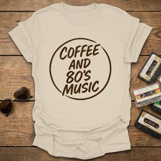 Beige t-shirt with 'Coffee and 80's Music' text on a wooden surface with cassette tapes and sunglasses.