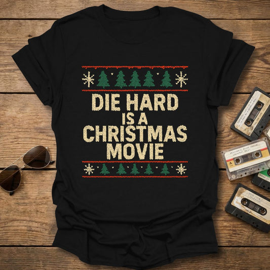 Black t-shirt with sweater pattern and 'Die Hard is a Christmas Movie' text on a wooden surface with cassettes and sunglasses.