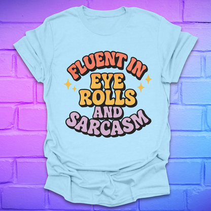 Light blue t-shirt with colorful text, 'fluent in eye rolls and sarcasm' design on a purple brick wall background