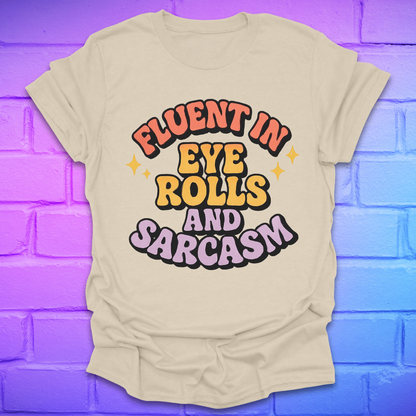 Beige t-shirt with colorful text, 'fluent in eye rolls and sarcasm' design on a purple brick wall background