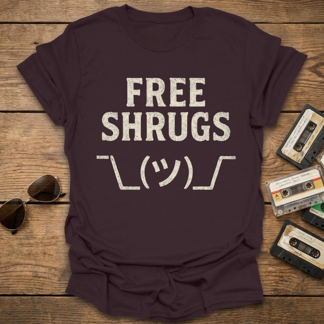 Brown t-shirt with 'FREE SHRUGS' text on a wooden surface with sunglasses and cassettes.
