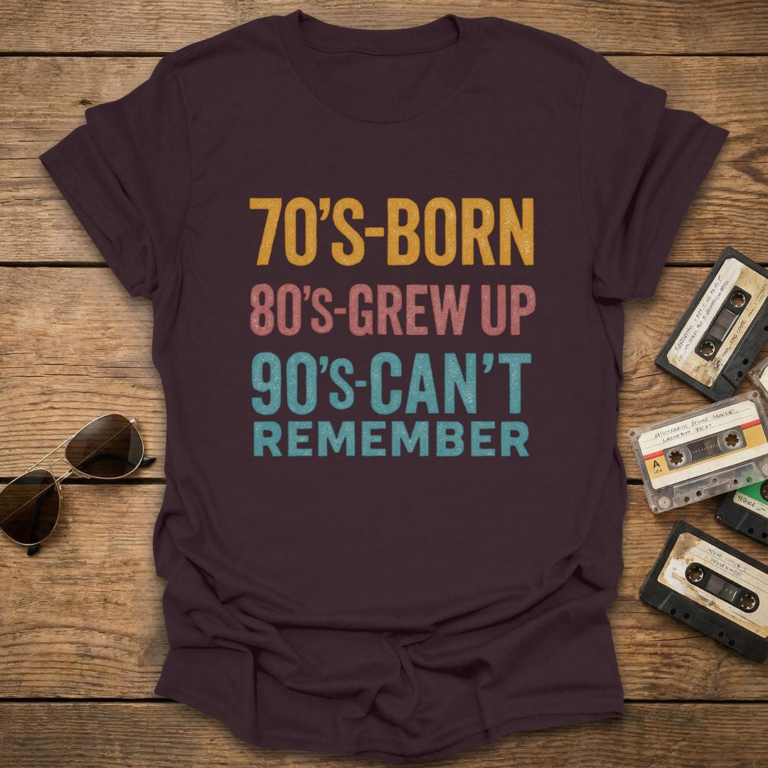 Brown t-shirt with text '70's-born 80's-grew up 90's-can't remember' on a wooden surface with vintage items.