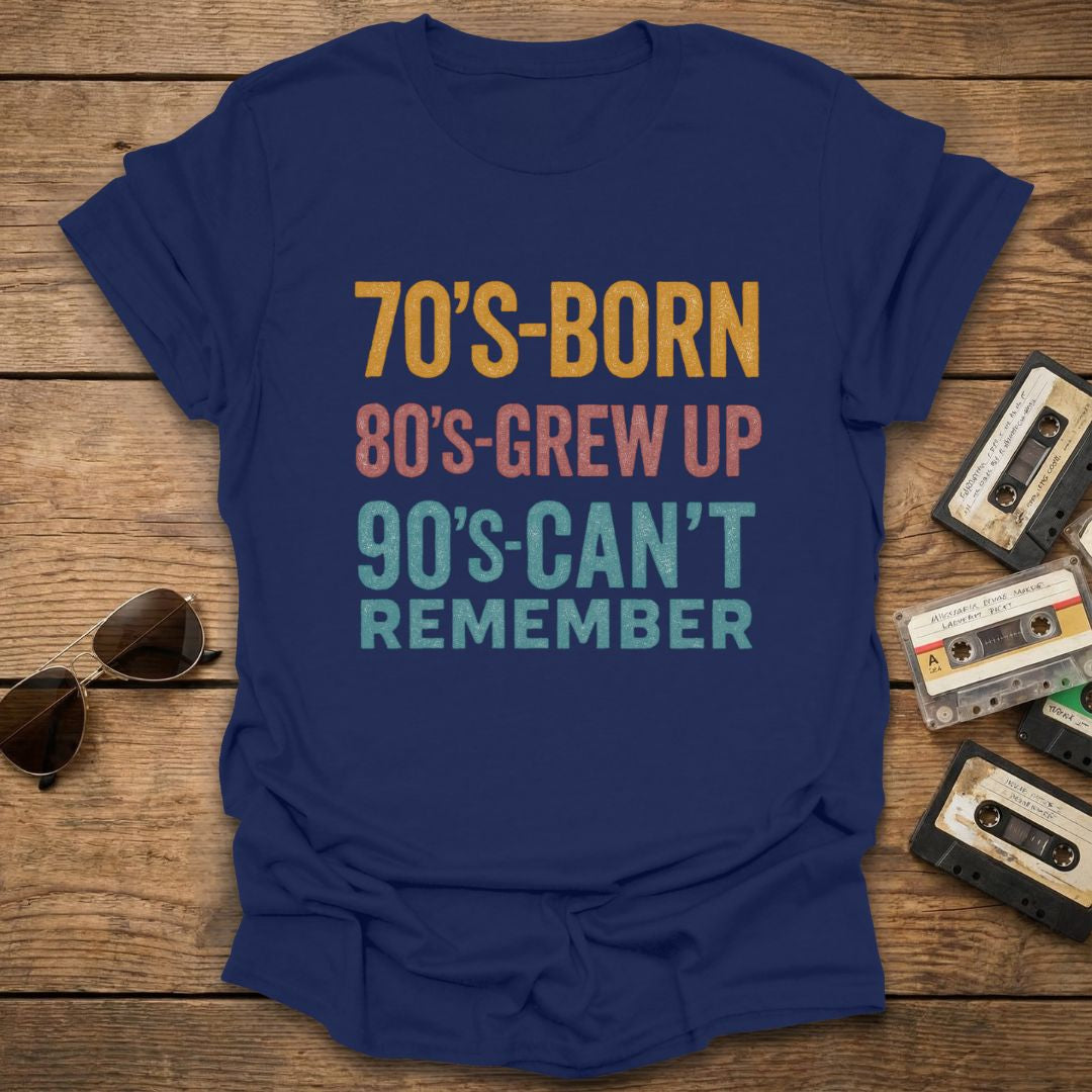 Navy blue t-shirt with text '70's-born 80's-grew up 90's-can't remember' on a wooden surface with sunglasses and cassettes.