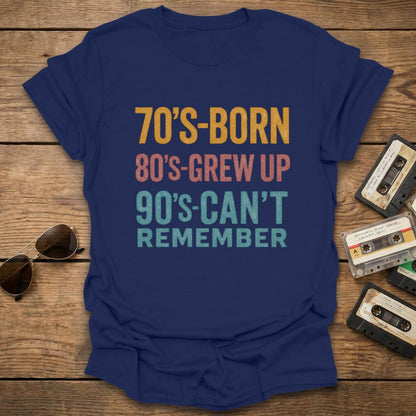 Navy blue t-shirt with text '70's-born 80's-grew up 90's-can't remember' on a wooden surface with sunglasses and cassettes.