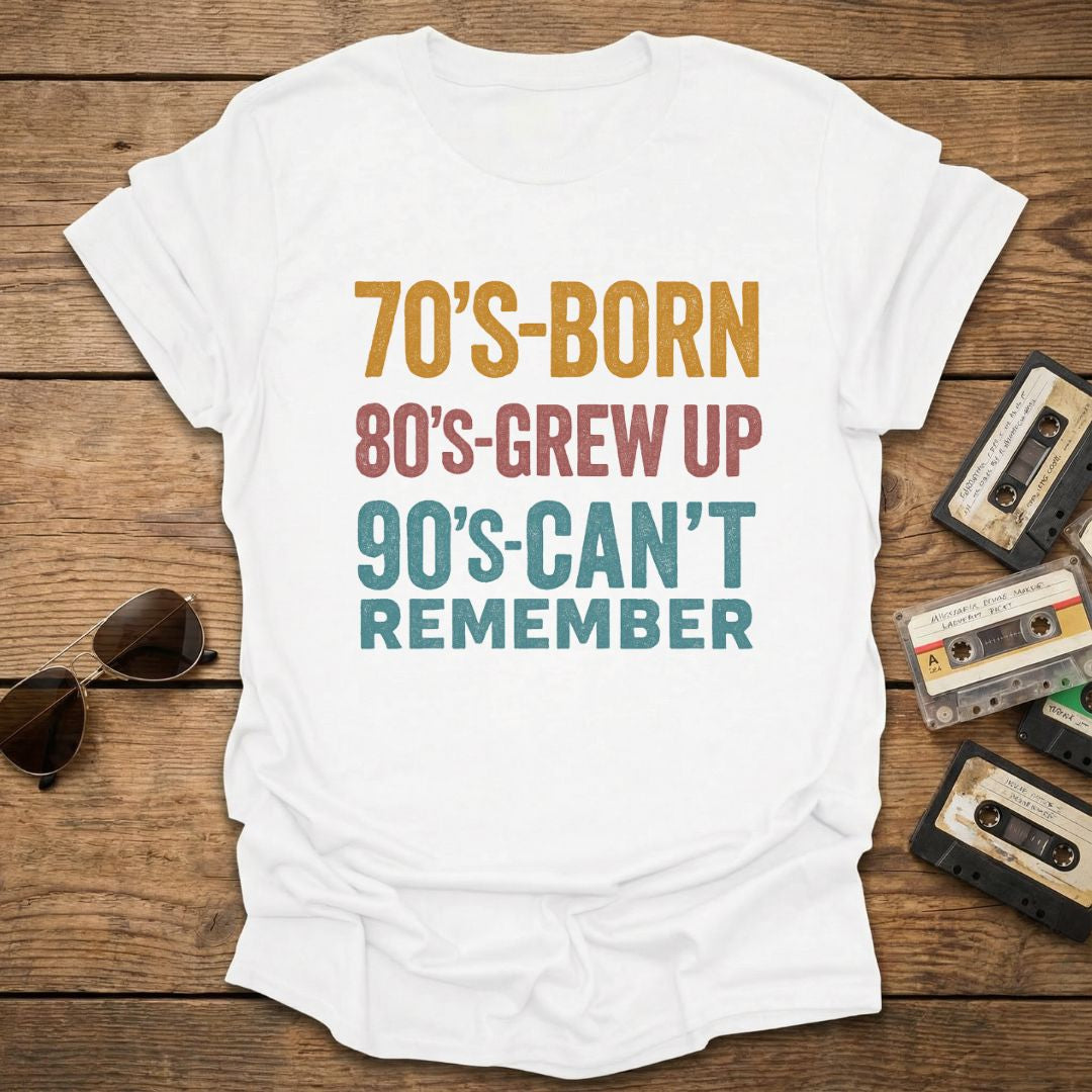 White t-shirt with text '70's-born, 80's-grew up, 90's-can't remember' on a wooden surface with vintage items.