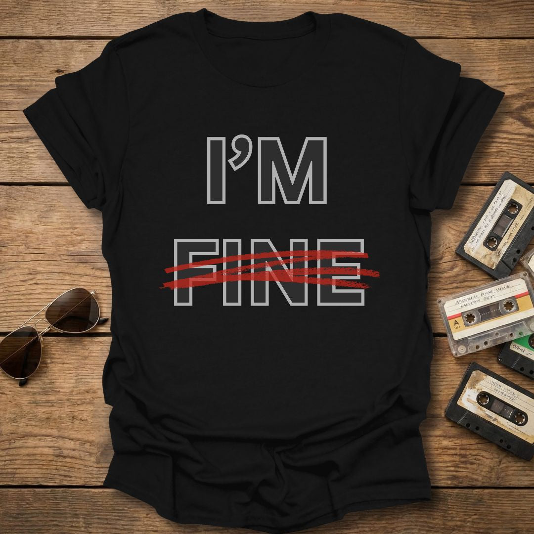 Black t-shirt with 'I'm Fine' with fine crossed out in red text on a wooden surface with sunglasses and cassettes.