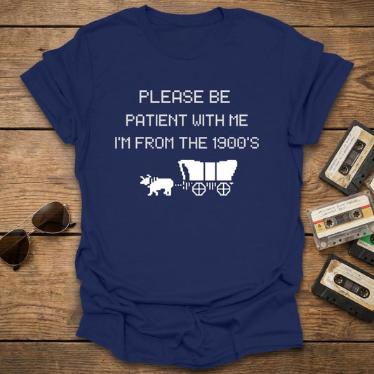 Navy blue t-shirt with text , 'please be patient with me im from the 1900s' and graphics on a wooden surface with vintage items.