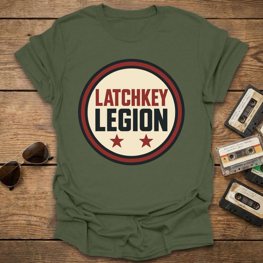 Green t-shirt with 'LATCHKEY LEGION' design on a wooden surface with sunglasses and cassettes.