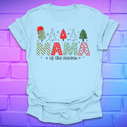 Light blue t-shirt with 'Mama of the season' text and Christmas tree graphics on a purple brick wall background.