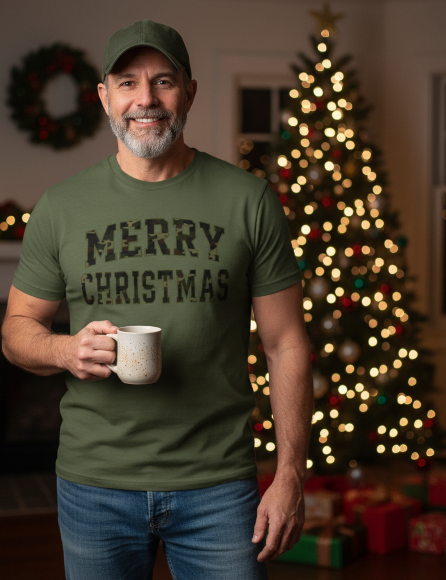 Man wearing a green 'Merry Christmas' t-shirt holding a mug in front of a decorated Christmas tree.