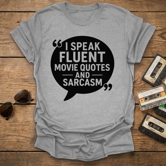 Gray t-shirt with 'I speak fluent movie quotes and sarcasm' text on a wooden surface with sunglasses and cassettes.