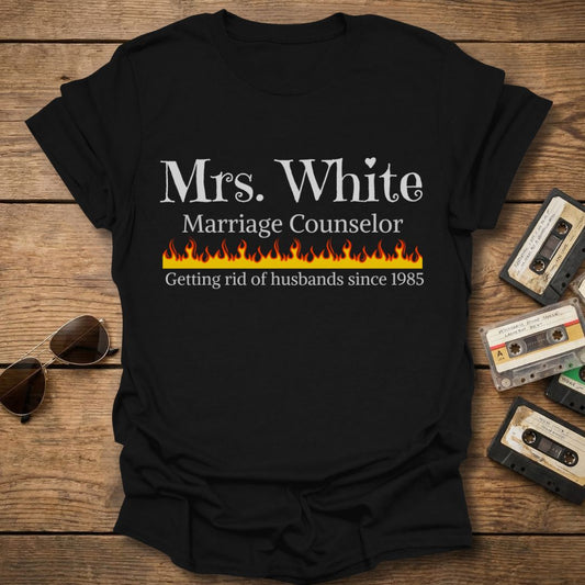 Black t-shirt with 'Mrs. White Marriage Counselor' text on a wooden surface with vintage tapes and sunglasses.