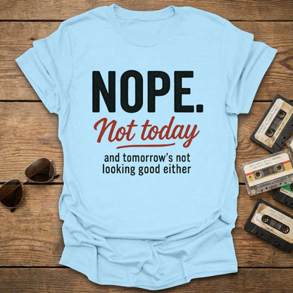 Light blue t-shirt with text 'NOPE. Not today and tomorrow's not looking good either' on a wooden surface with vintage cassettes and sunglasses.