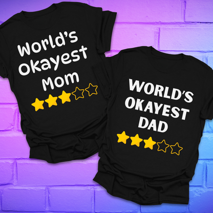 Two black t-shirts with 'World's Okayest Mom' and 'World's Okayest Dad' text on a purple brick wall background.