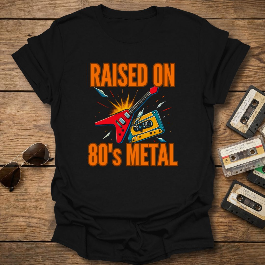 Black t-shirt with 'Raised on 80's Metal' design featuring a guitar and cassette tapes on a wooden surface with vintage cassettes and sunglasses.