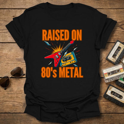 Black t-shirt with 'Raised on 80's Metal' design featuring a guitar and cassette tapes on a wooden surface with vintage cassettes and sunglasses.