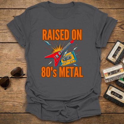 Gray t-shirt with 'Raised on 80's Metal' graphic design on a wooden surface with cassette tapes and sunglasses.