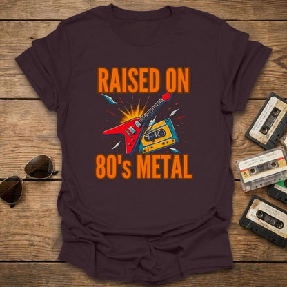 Brown T-shirt with 'Raised on 80's Metal' design on a wooden surface with cassette tapes and sunglasses.