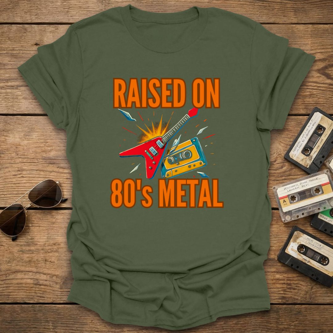 Green t-shirt with 'Raised on 80's Metal' design on a wooden surface with cassette tapes and sunglasses.