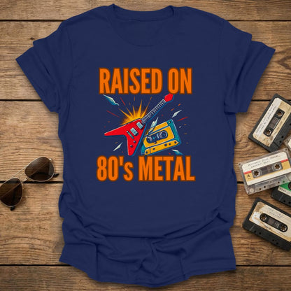 Blue t-shirt with 'Raised on 80's Metal' design on a wooden surface with cassette tapes and sunglasses.