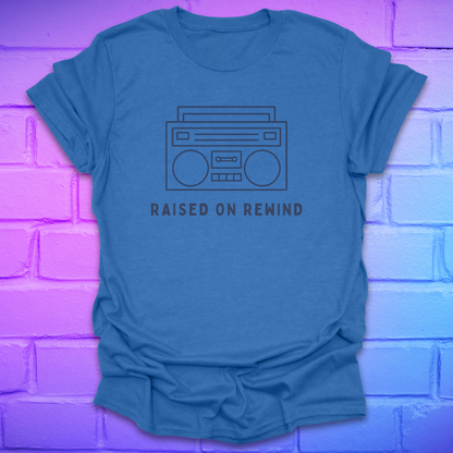 Heather royal t-shirt with a graphic and 'Raised on Rewind' text against a purple brick wall.