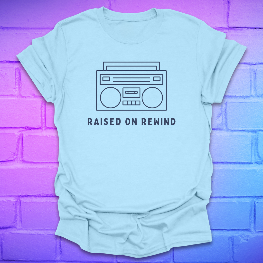 Light blue t-shirt with a graphic of a boombox and text 'Raised on Rewind' against a purple brick wall.