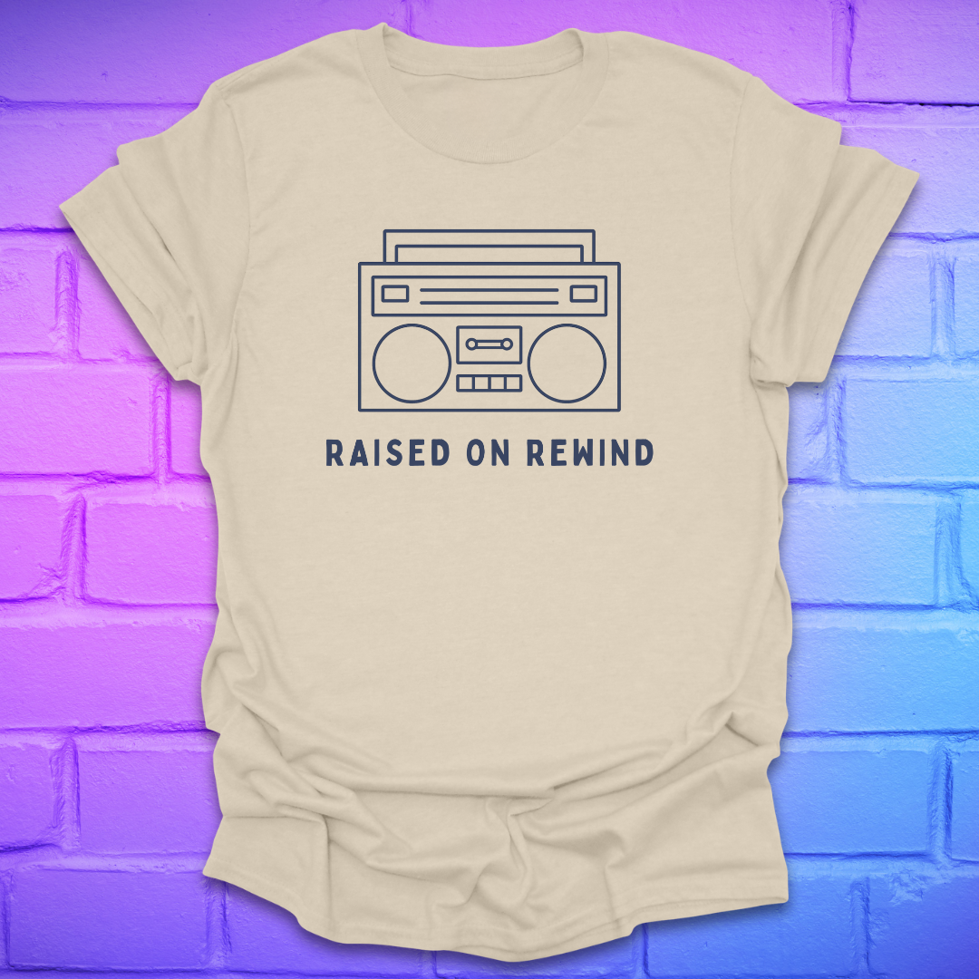Beige t-shirt with a graphic of a boombox and 'Raised on Rewind' text on a blue and purple brick wall background.
