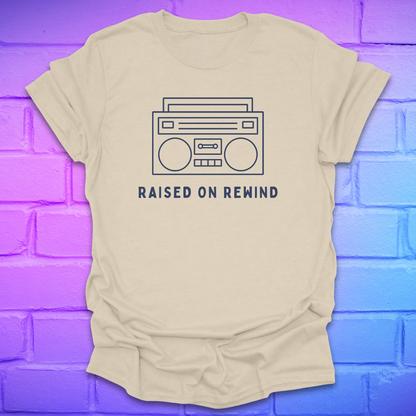 Beige t-shirt with a graphic of a boombox and 'Raised on Rewind' text on a blue and purple brick wall background.