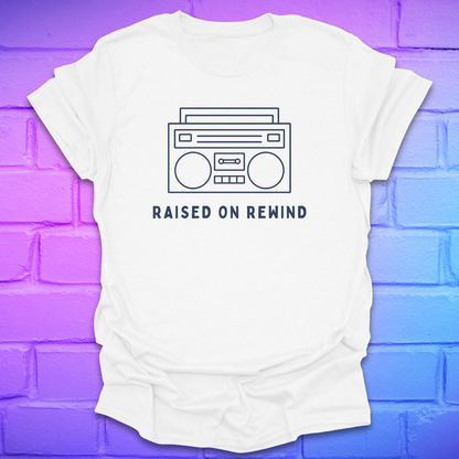 White t-shirt with a boombox graphic and 'Raised on Rewind' text on a gradient purple to blue brick wall.