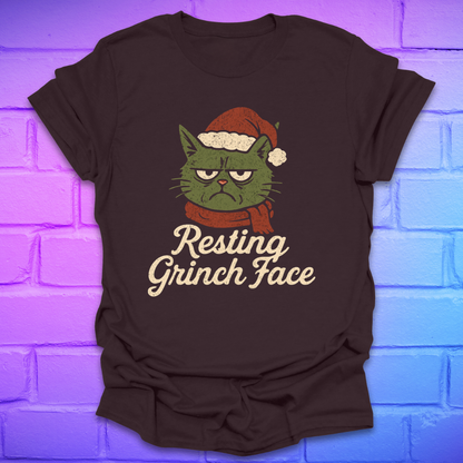 Brown t-shirt with a cartoon cat wearing a Santa hat and scarf, text 'Resting Grinch Face' on a brick wall background.