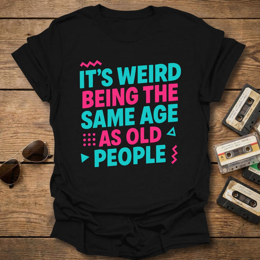 Black t-shirt with colorful text 'it's weird being the same age as old people', on a wooden surface with vintage cassettes and sunglasses.