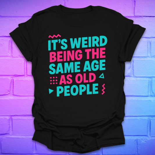 Black t-shirt with colorful text, 'it's weird being the same age as old people' on a brick wall background