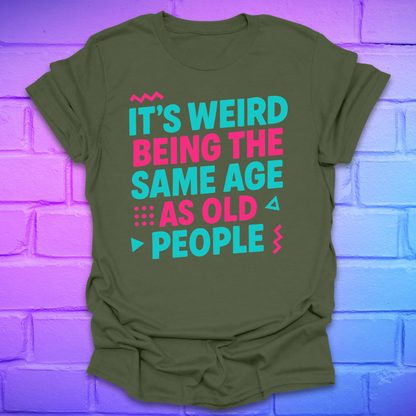 Green t-shirt with colorful text, 'it's weird being the same age as old people' on a brick wall background