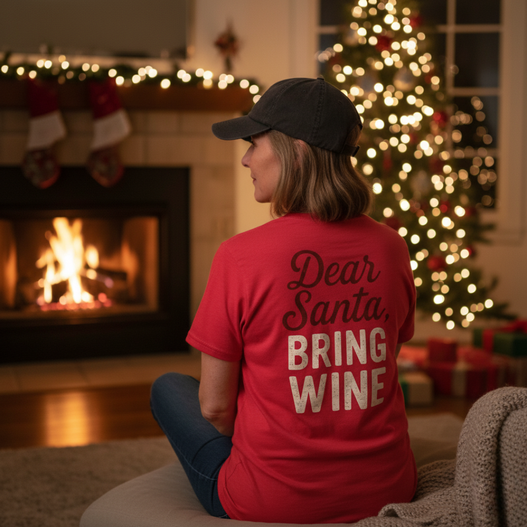 Person wearing a red shirt with 'Dear Santa, Bring Wine' text, on the back, in a festive room with a fireplace and Christmas tree.