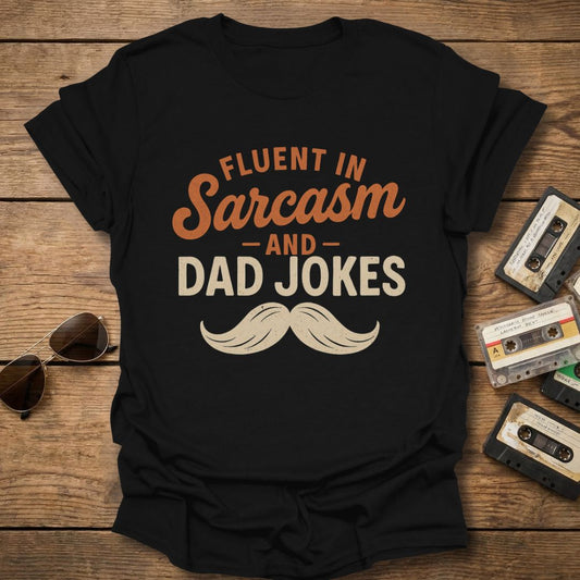 Black t-shirt with 'Fluent in Sarcasm and Dad Jokes' text on a wooden surface with cassettes and sunglasses.