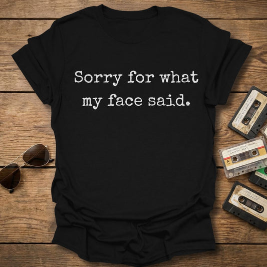 Black t-shirt with 'Sorry for what my face said' text on a wooden surface with sunglasses and cassettes.