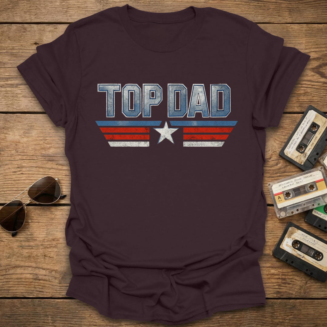 Brown t-shirt with 'Top Dad' graphic on a wooden surface with sunglasses and cassettes.