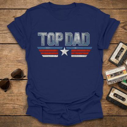 Blue t-shirt with 'TOP DAD' print on a wooden surface with sunglasses and cassettes.